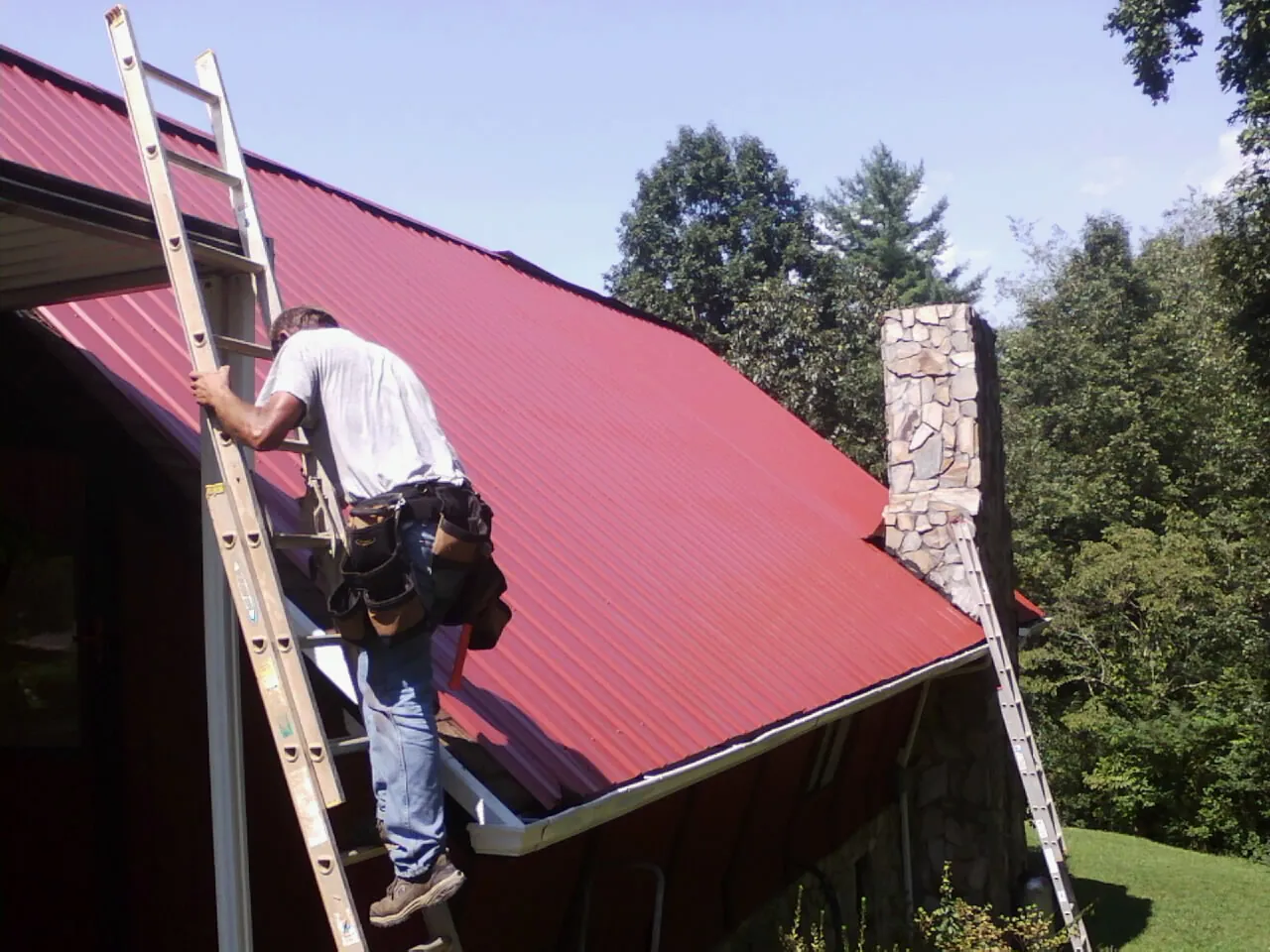 Roofer on a ladder beside a red metal roof and stone chimney — Soffit & Fascia Repair by Roofing Brothers in Hookstown